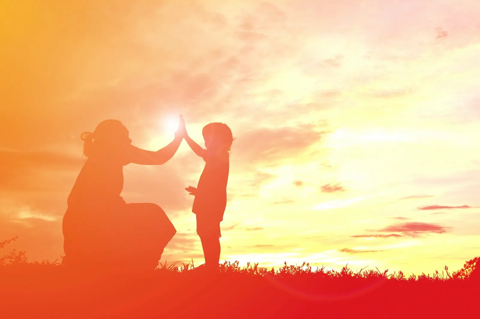 Silhouettes au coucher du soleil : une mère accroupie et un jeune enfant debout, paume contre paume, le soleil entre leurs mains.